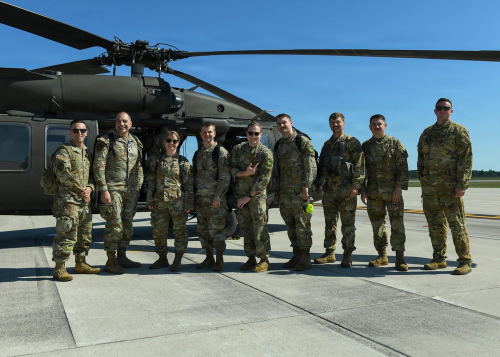 A group of nine military personnel in OCP (Operational Camouflage Pattern) uniforms pose for a group photo on a tarmac in front of a UH-60 Black Hawk helicopter. The setting appears to be a military airfield on a clear, sunny day. The group includes both male and female service members, suggesting a mixed unit or crew.