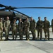 A group of nine military personnel in OCP (Operational Camouflage Pattern) uniforms pose for a group photo on a tarmac in front of a UH-60 Black Hawk helicopter. The setting appears to be a military airfield on a clear, sunny day. The group includes both male and female service members, suggesting a mixed unit or crew.