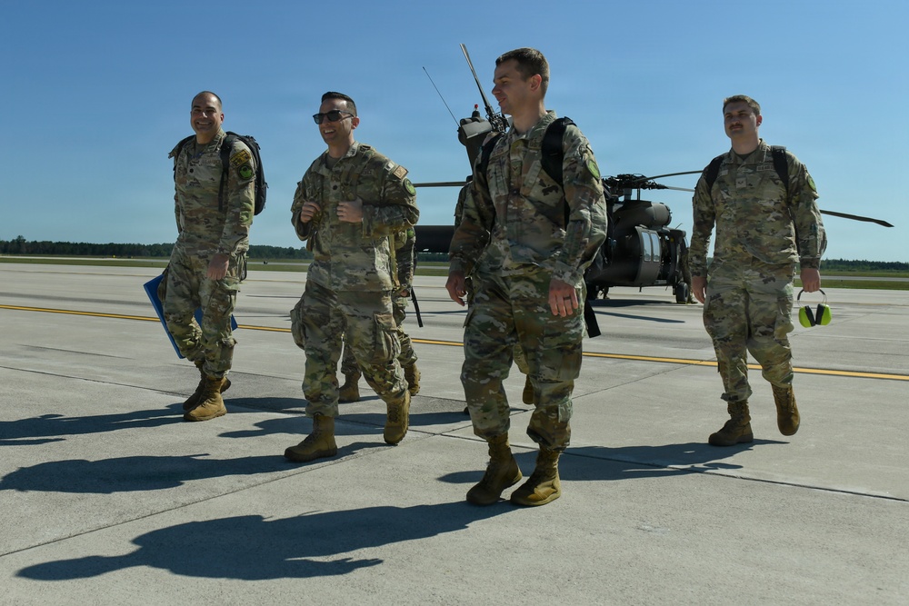Four military personnel in OCP (Operational Camouflage Pattern) uniforms are walking across a flight line tarmac, appearing relaxed and in good spirits. A military helicopter, consistent with a UH-60 Black Hawk, is visible in the background. The setting appears to be a military airfield under clear blue sky conditions.