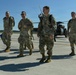 Four military personnel in OCP (Operational Camouflage Pattern) uniforms are walking across a flight line tarmac, appearing relaxed and in good spirits. A military helicopter, consistent with a UH-60 Black Hawk, is visible in the background. The setting appears to be a military airfield under clear blue sky conditions.