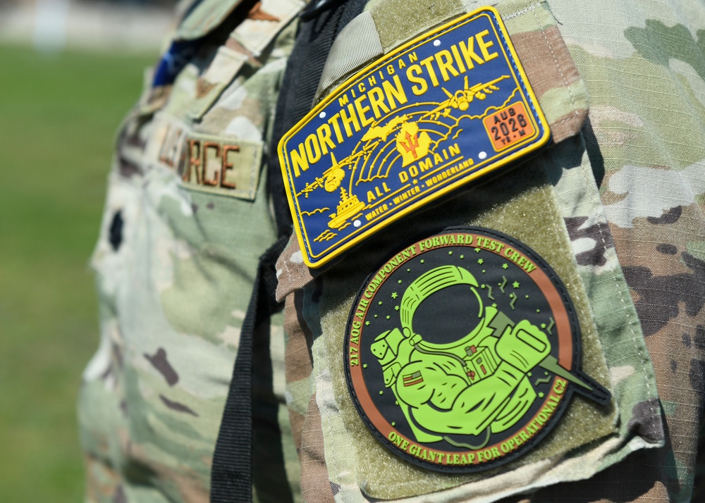 Close-up photograph of a military uniform sleeve displaying two tactical patches. The top patch is for 'Michigan Northern Strike' exercise (AUG 2028), an All Domain training event with the tagline 'Water, Winter, Wonderland.' The bottom patch belongs to the '217 AOG Air Component Forward Test Crew' with the motto 'One Giant Leap for Operational C2,' featuring an astronaut illustration.