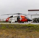 A U.S. Coast Guard MH-60 Jayhawk helicopter (tail number 6034) is parked on a tarmac near a large hangar facility. At least one crew member in an orange flight suit is visible near the aircraft's cabin door. A military tactical vehicle, appearing to be an HIMARS or similar wheeled launcher system, is parked nearby on the same tarmac.