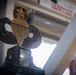The image shows a military trophy or award featuring a large paratrooper/airborne badge design with wings and a star, mounted on a dark pedestal base with an engraved plaque. The award appears to be the 'SSM Christopher L. Callan International Jumpmaster Award' based on partially visible text on the base. The trophy is photographed in an indoor setting with structural beams and a sign visible in the background.