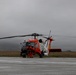 A U.S. Coast Guard helicopter, identifiable by its distinctive orange and white livery, is parked on a tarmac under overcast, foggy conditions. Two personnel dressed in orange flight suits are standing near the front of the aircraft, appearing to conduct pre-flight or post-flight checks. The scene suggests an operational readiness or search-and-rescue context at a coastal airfield.