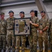 A group of military personnel pose together during what appears to be an awards ceremony inside a hangar or large indoor facility. Several individuals wearing blue berets are holding a framed award display containing medals and ribbons, suggesting a first-place team achievement. The group includes both U.S. Army soldiers and what appear to be allied military personnel in similar multicam uniforms.