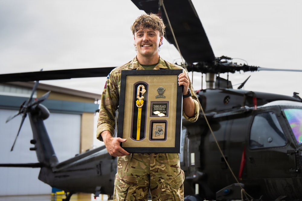 A smiling military personnel in MultiCam/MTP camouflage uniform holds a framed award plaque in front of military helicopters, likely UH-60 Black Hawks. The framed display contains what appears to be a yellow-handled tool or instrument, an Airborne badge/insignia, and a plaque reading 'Third Place Individual' with a partially visible date (2026). The setting appears to be a military airfield or flight line.