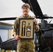 A smiling military personnel in MultiCam/MTP camouflage uniform holds a framed award plaque in front of military helicopters, likely UH-60 Black Hawks. The framed display contains what appears to be a yellow-handled tool or instrument, an Airborne badge/insignia, and a plaque reading 'Third Place Individual' with a partially visible date (2026). The setting appears to be a military airfield or flight line.