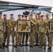 A group of approximately 12 military personnel, appearing to be a mix of U.S. Army and British Armed Forces members, pose together in front of a helicopter on a tarmac or hangar apron. They are holding two framed award plaques, suggesting a competition or achievement recognition ceremony. The group is smiling, indicating a celebratory occasion.