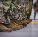 The image shows a row of military personnel standing at attention in formation, photographed from a low angle focusing on their boots and lower legs. All visible individuals are wearing matching MultiCam/OCP camouflage uniforms and tan/coyote brown combat boots. A single figure stands in the background, slightly out of focus, appearing to observe or oversee the formation.