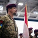 A military personnel member wearing a maroon beret and camouflage uniform stands at attention during what appears to be a formal military ceremony. The name tape on the uniform reads 'LE BOURDAIS' and the sleeve displays multiple patches including what appears to be a Canadian flag-related insignia and a Ukraine-related patch. A red and white flag (consistent with Canadian colors) is visible in the background alongside other personnel in maroon berets.