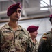 The image shows a group of military personnel in uniform, with a U.S. Army soldier named Bhattacharyya prominently in the foreground wearing a maroon beret and OCP (Operational Camouflage Pattern) uniform. A German Bundeswehr soldier is visible on the right, identifiable by the German flag patch and Flecktarn camouflage uniform. The setting appears to be an indoor military facility, suggesting a joint multinational military event or ceremony.