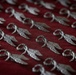 The image shows a large collection of identical silver metal insignia or badges arranged in rows on a red velvet surface. Each badge features a wreath or laurel ring with wings extending from both sides, suggesting a military, aviation, or ceremonial context. The items appear to be newly manufactured or prepared for distribution.
