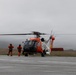 A U.S. Coast Guard MH-60 Jayhawk helicopter, numbered 34, sits on a tarmac with its rotors spinning. Two crew members dressed in orange survival suits and helmets are approaching the aircraft, apparently preparing to board. The scene appears to be at a remote coastal airfield under overcast, foggy conditions.