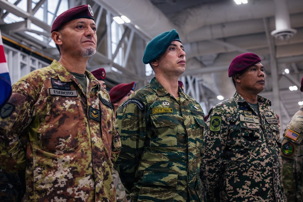 The image shows military personnel from multiple nations standing at attention, likely during a multinational military ceremony or event held indoors in what appears to be a hangar or large military facility. Three prominent soldiers are visible in the foreground wearing distinct national uniforms and insignia. The gathering appears to be a joint international military exercise or formal ceremony.