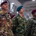 The image shows military personnel from multiple nations standing at attention, likely during a multinational military ceremony or event held indoors in what appears to be a hangar or large military facility. Three prominent soldiers are visible in the foreground wearing distinct national uniforms and insignia. The gathering appears to be a joint international military exercise or formal ceremony.