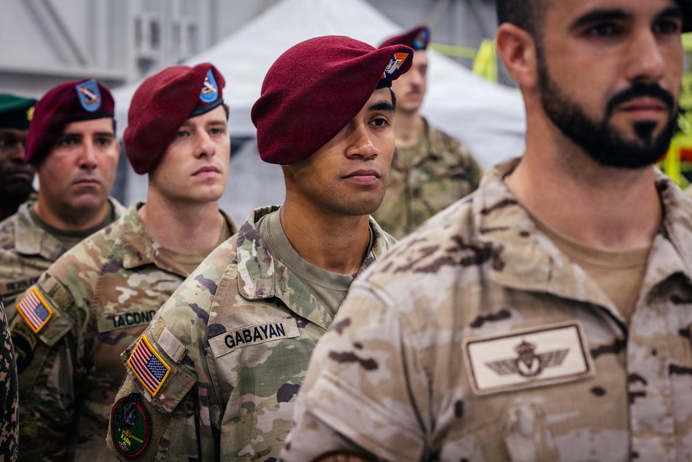 A group of military personnel in formation, wearing maroon berets indicating airborne/paratrooper units. At least two different national uniforms are visible, suggesting a multinational military exercise or ceremony. The soldiers appear to be at attention or standing in a formal assembly.