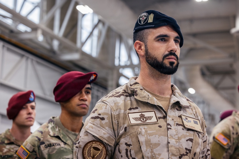 The image shows military personnel from multiple nations standing in formation inside what appears to be a hangar or large military facility. The foreground subject wears a Spanish military uniform with a black beret bearing a parachutist badge, identified by the name tape 'DELGADO' and an 'Ejército del Aire y del Espacio' patch. Behind him are US Army soldiers wearing maroon berets and MultiCam uniforms, one identified by the name tape 'GABAYAN'.