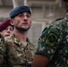 The image shows multiple military personnel from what appear to be different nations gathered together, likely at a joint military event or ceremony held inside a large facility. The central figure wears British MTP (Multi-Terrain Pattern) camouflage and a blue beret, consistent with Royal Air Force dress. Other visible personnel wear a maroon beret and a distinct green digital camouflage uniform with patches suggesting a different national military.