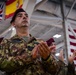 A military ceremony taking place indoors, likely in a hangar or large military facility. The central figure is an Italian Army officer wearing a maroon beret and camouflage uniform with 'ESERCITO' (Italian for 'Army') name tape, applauding during the event. Multiple national flags are visible in the background, indicating a multinational military gathering.