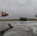 A U.S. Coast Guard helicopter, painted in its distinctive orange and white livery, is hovering or landing on a wet tarmac during overcast, rainy conditions. A soldier in camouflage uniform is signaling the aircraft with raised hands, while a military tactical truck is parked in the background. The scene appears to depict a joint military and Coast Guard operation or exercise.