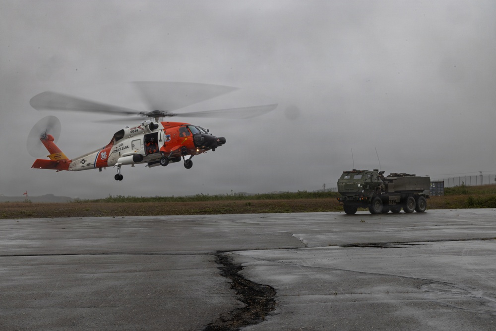 A U.S. Coast Guard helicopter, painted in its distinctive orange and white livery, is captured hovering or taking off from a wet tarmac under overcast, stormy conditions. A military tactical truck is parked nearby on the same tarmac. The scene suggests an emergency response or joint military-Coast Guard operation during adverse weather.