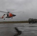 A U.S. Coast Guard helicopter, painted in its distinctive orange and white livery, is captured hovering or taking off from a wet tarmac under overcast, stormy conditions. A military tactical truck is parked nearby on the same tarmac. The scene suggests an emergency response or joint military-Coast Guard operation during adverse weather.