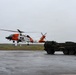 A U.S. Coast Guard helicopter, marked with the distinctive orange and white color scheme and tail number 6034, is hovering or landing on a wet tarmac in overcast, rainy conditions. A large military tactical truck, consistent with a HEMTT (Heavy Expanded Mobility Tactical Truck) or similar vehicle, is parked nearby on the same surface. The scene appears to depict a joint military or interagency operation at a remote airfield.
