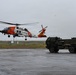 A U.S. Coast Guard MH-60 Jayhawk helicopter (tail number 6034) is hovering or taking off from a wet tarmac in overcast, rainy conditions. Nearby on the tarmac is a military ground vehicle that appears to be an M270 or HIMARS-type rocket artillery launcher system on a heavy tactical truck. The scene suggests a joint military exercise or coordinated operation between U.S. Coast Guard and Army assets.