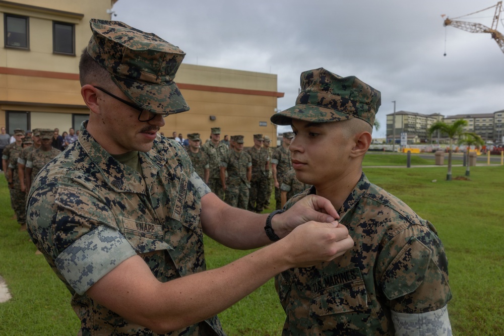 A U.S. Marine identified by the name tape 'KNAPP' is pinning a medal or insignia onto another Marine during what appears to be a formal military awards ceremony. The event is taking place outdoors on a military installation, with a formation of Marines standing at attention in the background. The setting appears to be a base in a tropical or Pacific region, suggested by the palm trees and building architecture.