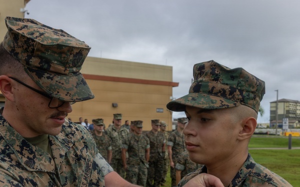 A U.S. Marine identified by the name tape 'KNAPP' is pinning a medal or insignia onto another Marine during what appears to be a formal military awards ceremony. The event is taking place outdoors on a military installation, with a formation of Marines standing at attention in the background. The setting appears to be a base in a tropical or Pacific region, suggested by the palm trees and building architecture.