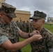 A U.S. Marine identified by the name tape 'KNAPP' is pinning a medal or insignia onto another Marine during what appears to be a formal military awards ceremony. The event is taking place outdoors on a military installation, with a formation of Marines standing at attention in the background. The setting appears to be a base in a tropical or Pacific region, suggested by the palm trees and building architecture.
