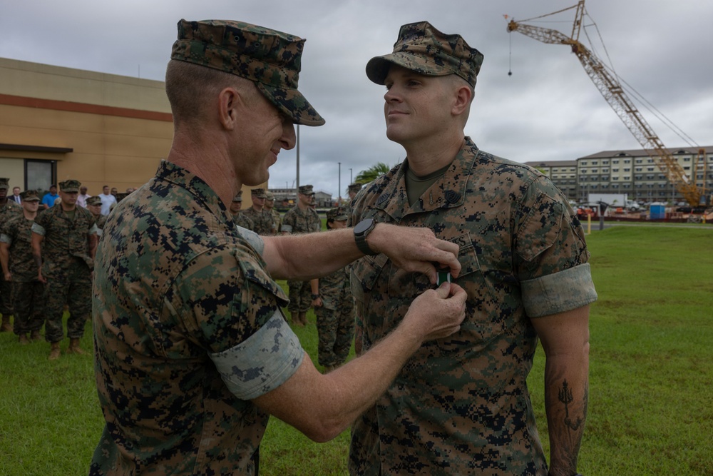 Two U.S. Marines in MARPAT woodland camouflage uniforms are engaged in a military decoration ceremony outdoors. One Marine is pinning a medal or award onto the chest of another Marine while a formation of service members observes in the background. The setting appears to be a military installation, likely in a tropical or Pacific region based on the vegetation and construction visible.