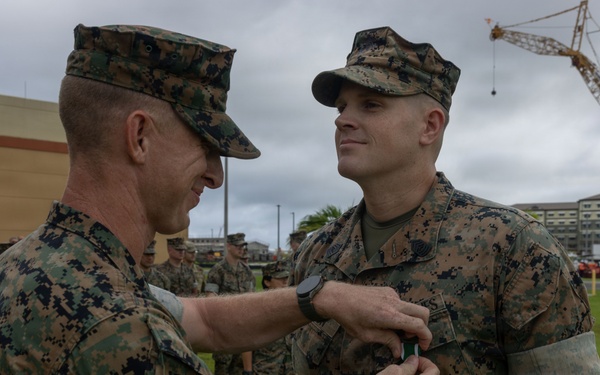 Two U.S. Marines in MARPAT woodland camouflage uniforms are engaged in a military decoration ceremony outdoors. One Marine is pinning a medal or award onto the chest of another Marine while a formation of service members observes in the background. The setting appears to be a military installation, likely in a tropical or Pacific region based on the vegetation and construction visible.