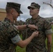 Two U.S. Marines in MARPAT woodland camouflage uniforms are engaged in a military decoration ceremony outdoors. One Marine is pinning a medal or award onto the chest of another Marine while a formation of service members observes in the background. The setting appears to be a military installation, likely in a tropical or Pacific region based on the vegetation and construction visible.