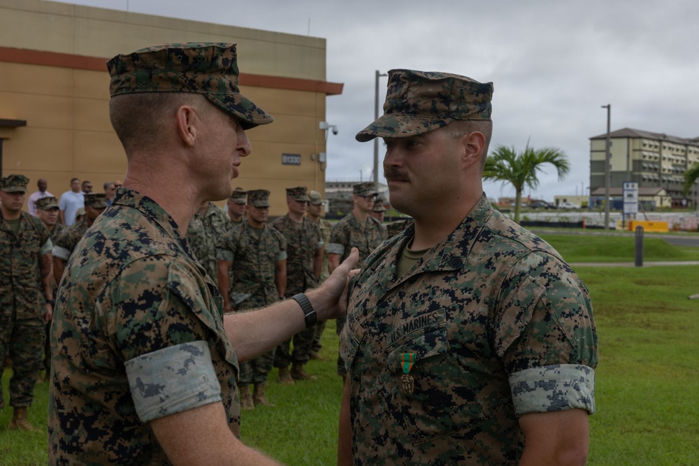 A U.S. Marine Corps award ceremony is taking place outdoors at what appears to be a military installation. A senior Marine is pinning or presenting a medal to another Marine who is wearing a ribbon/medal on his chest, while a formation of Marines stands at attention in the background. The setting appears to be a tropical or subtropical location based on the palm trees and building architecture visible.