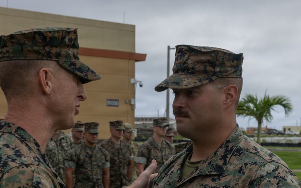 A U.S. Marine Corps award ceremony is taking place outdoors at what appears to be a military installation. A senior Marine is pinning or presenting a medal to another Marine who is wearing a ribbon/medal on his chest, while a formation of Marines stands at attention in the background. The setting appears to be a tropical or subtropical location based on the palm trees and building architecture visible.