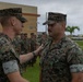 A U.S. Marine Corps award ceremony is taking place outdoors at what appears to be a military installation. A senior Marine is pinning or presenting a medal to another Marine who is wearing a ribbon/medal on his chest, while a formation of Marines stands at attention in the background. The setting appears to be a tropical or subtropical location based on the palm trees and building architecture visible.