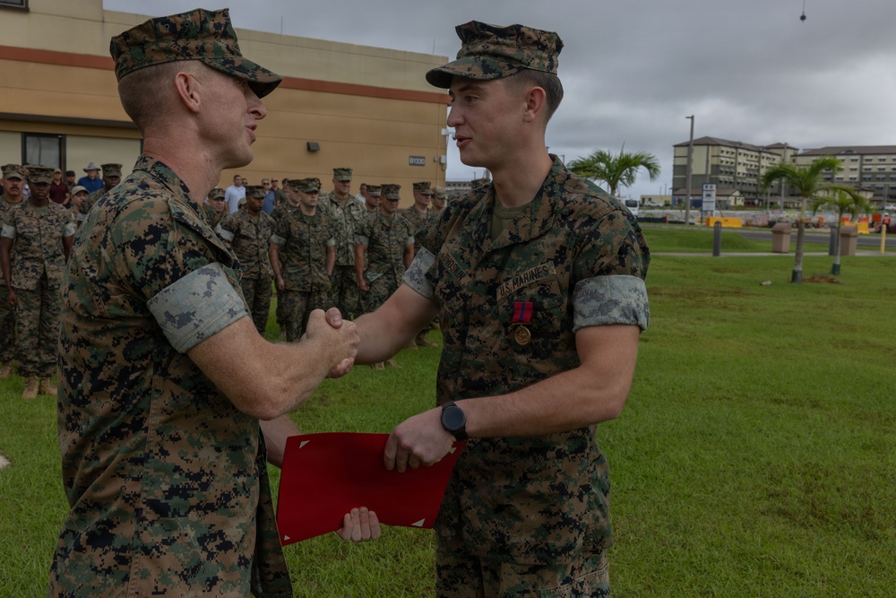 Two U.S. Marines in MARPAT camouflage uniforms are shaking hands during what appears to be a formal military award or recognition ceremony. The Marine on the right is wearing a medal and ribbon on his chest and holding a red folder, suggesting he is the recipient of a commendation. A formation of Marines stands in the background on a grassy area near military base buildings.