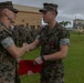 Two U.S. Marines in MARPAT camouflage uniforms are shaking hands during what appears to be a formal military award or recognition ceremony. The Marine on the right is wearing a medal and ribbon on his chest and holding a red folder, suggesting he is the recipient of a commendation. A formation of Marines stands in the background on a grassy area near military base buildings.