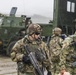 The image shows multiple soldiers in MultiCam/OCP camouflage uniforms operating in wet, rainy conditions near military vehicles. At least three personnel are visible in the foreground, armed and equipped with tactical gear, helmets, and body armor. In the background, olive-drab military vehicles — consistent with HIMARS or similar wheeled military platforms — are visible with amber warning lights active.