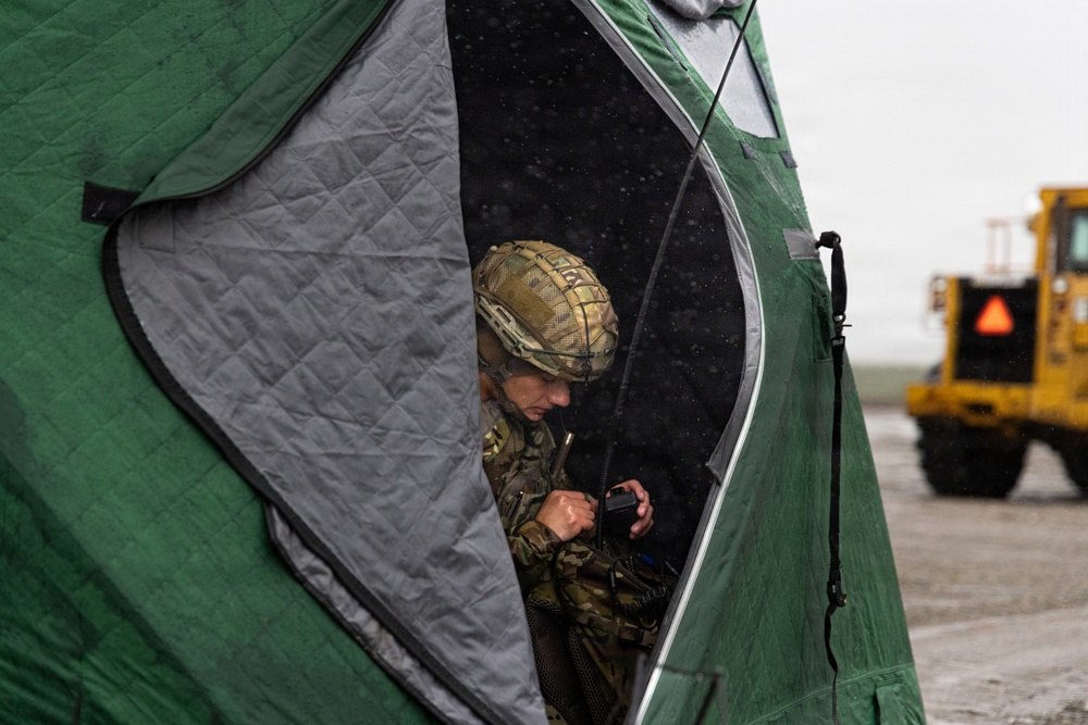 A military personnel wearing camouflage uniform and tactical helmet is sheltering inside a green insulated tent during what appears to be rainy or snowy weather conditions. The soldier is operating a handheld radio or communication device. A yellow heavy vehicle is visible in the background on what appears to be an airfield or open terrain.