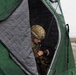 A military personnel wearing camouflage uniform and tactical helmet is sheltering inside a green insulated tent during what appears to be rainy or snowy weather conditions. The soldier is operating a handheld radio or communication device. A yellow heavy vehicle is visible in the background on what appears to be an airfield or open terrain.