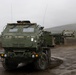 The image shows a U.S. Army M142 High Mobility Artillery Rocket System (HIMARS) vehicle positioned on wet, muddy terrain under overcast, foggy conditions. The vehicle is painted in olive drab green and bears crew name markings on its cab. A second similar vehicle is partially visible in the background, suggesting a unit-level field exercise or training operation.