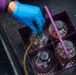 A gloved hand is inserting a hose or tube into one of four large glass jars or jugs contained in a purple plastic carrier. The jars appear to contain a brownish liquid, possibly being filled or sampled. The setting appears to be an outdoor or industrial environment with a wet, dark surface.