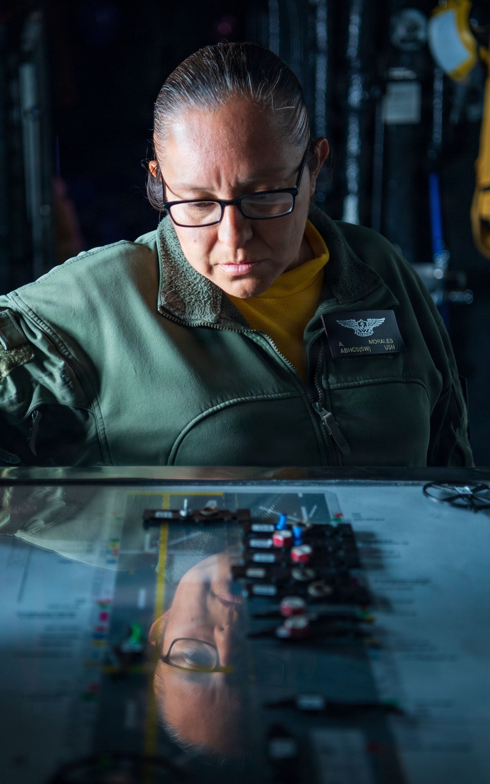A U.S. Navy service member identified by name tag as 'A. Morales, ABHCS(SW), USN' is focused on working at a glass-topped table or display board containing what appear to be colored markers or tokens arranged on a diagram. The individual is wearing a green flight deck jacket with a naval aviation warfare insignia patch and black-rimmed glasses. The reflective glass surface creates a mirror image of the person and the items on the table below.
