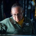 A U.S. Navy service member identified by name tag as 'A. Morales, ABHCS(SW), USN' is focused on working at a glass-topped table or display board containing what appear to be colored markers or tokens arranged on a diagram. The individual is wearing a green flight deck jacket with a naval aviation warfare insignia patch and black-rimmed glasses. The reflective glass surface creates a mirror image of the person and the items on the table below.