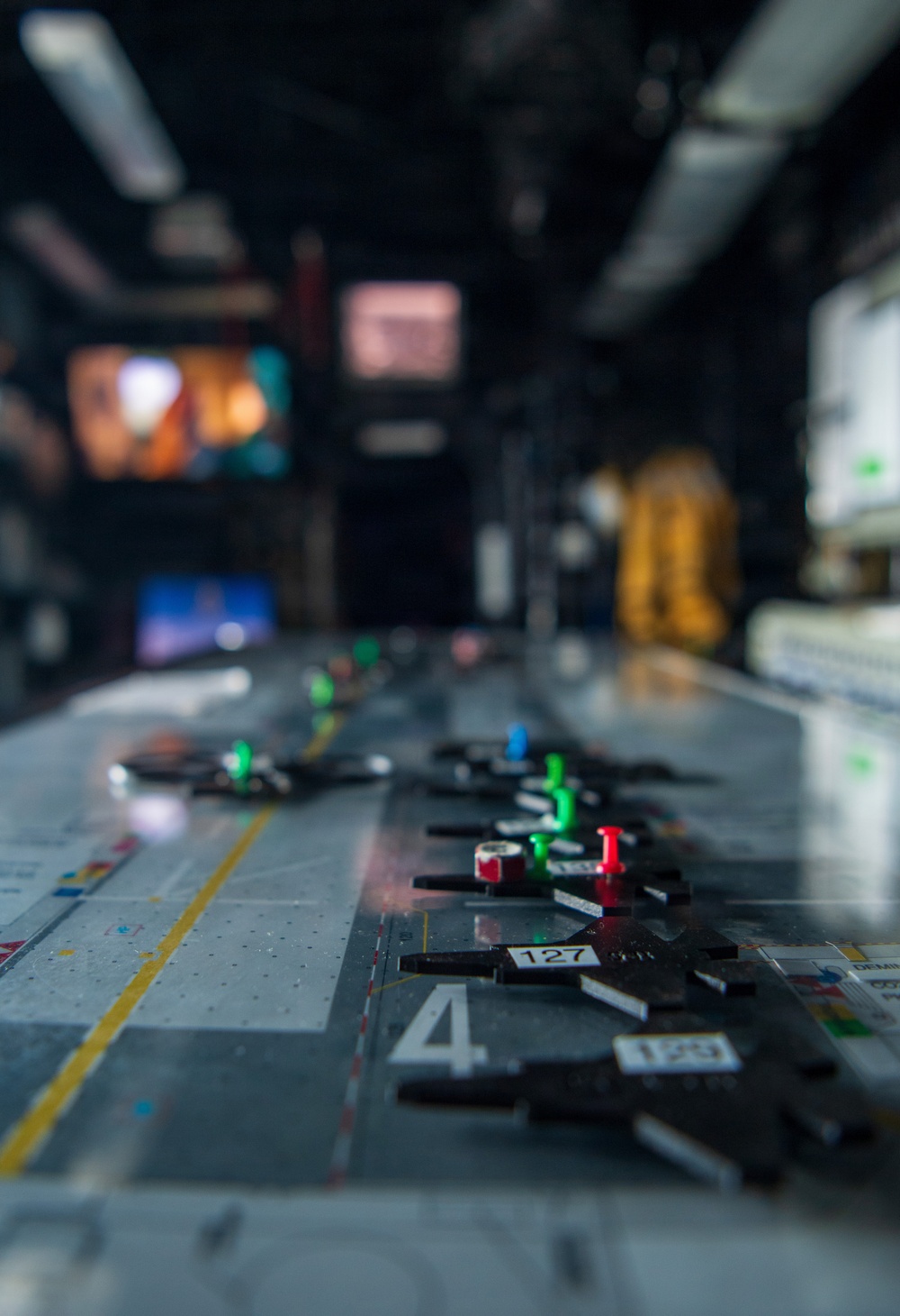 The image shows a close-up of what appears to be a naval aircraft carrier flight deck operations board or 'Ouija board,' used to track aircraft positions and movements. Small black aircraft-shaped tokens are arranged on a gray surface marked with numbers including '4,' '127,' and '133,' along with colored indicator pins (green, red, blue). The background is blurred but shows a dimly lit operations room with multiple display screens and at least one person in the background.