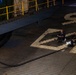 Two individuals dressed in dark clothing are seated on a corrugated metal deck surface, likely aboard a large vessel or at a dock facility. A large black hose or cable runs diagonally across the deck near them. The scene is lit by a single strong light source creating dramatic shadows, with yellow safety railings and green cargo containers visible in the background.