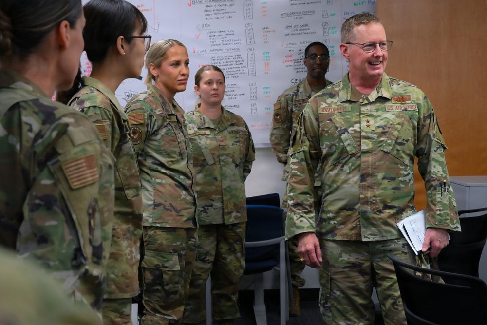 A group of U.S. Air Force personnel in OCP (Operational Camouflage Pattern) uniforms are gathered in what appears to be a briefing or training room. A senior officer, identifiable by a star insignia and name tape reading 'BRADFIELD' on a U.S. Air Force uniform, appears to be addressing or interacting with the group. A whiteboard covered in handwritten notes and training topics is visible in the background.