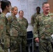 A group of U.S. Air Force personnel in OCP (Operational Camouflage Pattern) uniforms are gathered in what appears to be a briefing or training room. A senior officer, identifiable by a star insignia and name tape reading 'BRADFIELD' on a U.S. Air Force uniform, appears to be addressing or interacting with the group. A whiteboard covered in handwritten notes and training topics is visible in the background.